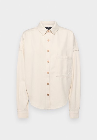 Camisa de manga larga color crema, con botones en la parte delantera, cuello, corte suelto, un bolsillo en el pecho y textura sutil. Acentos de botones en bronce.