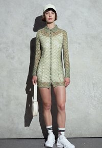 Overall verde transparente com padrão, mangas longas, frente de botões e calções. Combinado com um boné branco e uma mala, ténis brancos e meias às riscas.