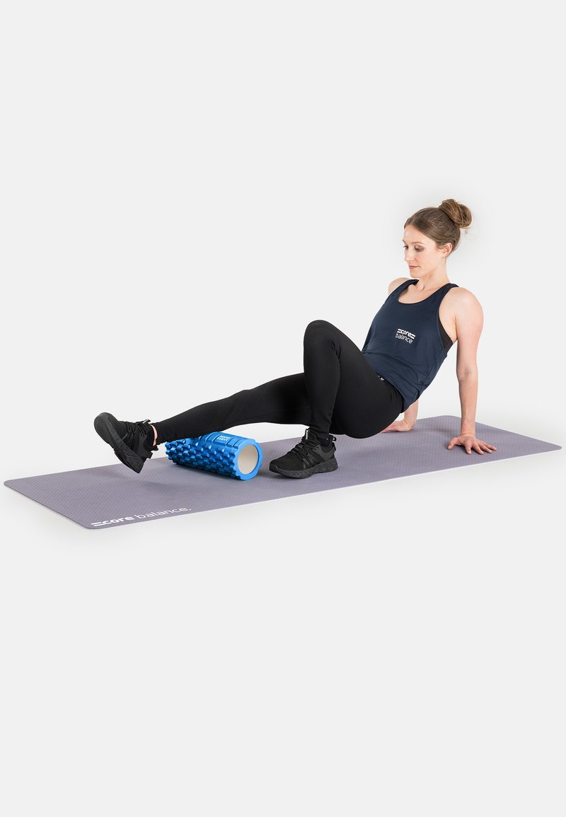 Blue foam roller with a textured surface on a grey exercise mat, used for physical therapy, with a person positioned beside it.