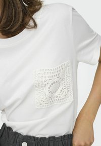 Hvid bomulds t-shirt med en tekstureret hvid hækle-lomme på venstre side, sat mod en lys baggrund. Korte ærmer, rund halsudskæring.
