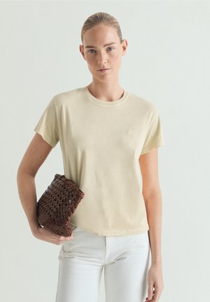 Kvinde med blondt hår i en beige kortærmet skjorte og hvide bukser, der holder en brun tekstureret clutch mod en ensartet lys baggrund.