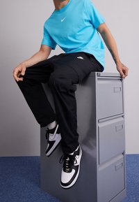 Persona con camiseta azul de Nike, pantalones cargo negros de Nike y zapatillas negro y blanco de Nike, sentada en un archivador gris.