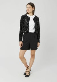 Veste bomber noire en sequins avec un col rond, coupe courte, associée à un short noir sur mesure et des talons noirs à brides.
