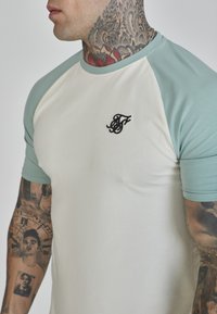 Man with detailed neck and arm tattoos wears a white and light green raglan sleeve shirt with a small black embroidered logo on chest.