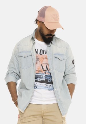 Homme portant une casquette rose, une chemise en jean clair sur un t-shirt graphique avec une voiture classique et des palmiers, et un short cargo beige, les mains dans les poches.
