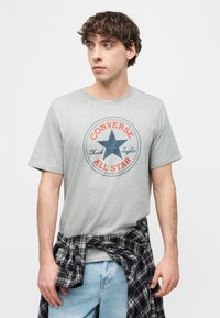 Νεαρός άνδρας που φοράει γκρί μπλουζάκι Converse All Star κ αι έχει δέσει ένα ασπρόμαυρο καρό πουκάμισο γύρω από τη μέση του, κοιτάζοντας προς τα αριστερά.