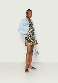 Jaqueta de ganga sobre um vestido preto com flores e uma fenda lateral. Sandálias com tira e uma pequena bolsa cinza. Design ajustado e casual.