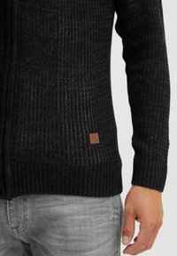 Sort strikket cardigan med en tekstureret ribbede design, der har et brunt lædermærke nederst til venstre. Slim fit med lange ærmer.