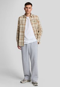 Camisa de cuadros en tonos marrón, naranja y crema, llevada sobre una camiseta blanca, combinada con unos pantalones de chándal grises y zapatillas deportivas con estampado.