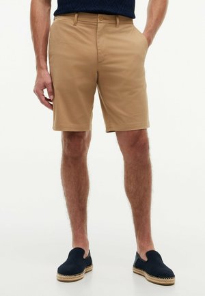 Mann trägt beige kniehohe Shorts mit Taschen und schwarze Slipper, steht vor weißem Hintergrund.