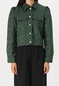 Grön jacquardkort jacka med texturerat blommönster, knäppning och två bröstfickor. Långa ärmar med knappade manschetter.