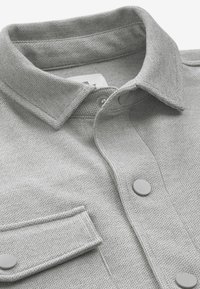 Camisa gris texturizada con cuello, que presenta un bolsillo en el pecho y botones grises redondos. La tela tiene un sutil patrón tejido.