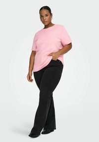 Różowy bawełniany t-shirt o luźnym kroju z krótkimi rękawami, zestawiony z czarnymi rozszerzanymi dżinsami. Modelka ma rękę w kieszeni i stoi lekko bokiem.