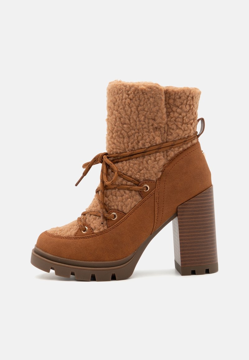 Bottine en daim marron avec talon épais empilé, doublure style peau de mouton, et lacets enveloppants noués sur le côté sur un fond blanc.