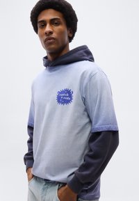 Νέος άνδρας με αφροχτένισμα που φορά στρωμένο μπλε φούτερ και μπλουζάκι με το λογότυπο "scotch & soda", τα χέρια στις τσέπες ανοιχτού τζιν.