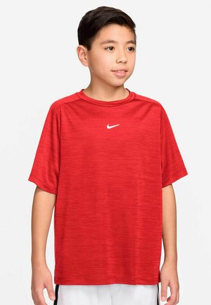 Ragazzo con capelli corti e scuri che indossa una maglietta sportiva rossa Nike e pantaloncini bianchi, rivolto leggermente verso destra su uno sfondo uniforme.