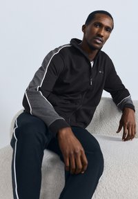 Čierna bunda so zapínaním na zips, sivé rukávy a biely bočný pásik. Mäkká látka, voľný strih, model sediaci na textúrovanom svetlom povrchu.