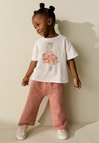Tricou roz deschis cu o ilustrație cu model floral, asortat cu pantaloni roz deschis, largi și texturizați, și adidași albi.