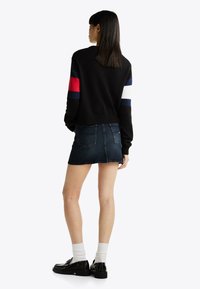 Maglione nero con strisce multicolori; abbinato a una minigonna in denim. Modella indossa scarpe nere e calzini bianchi, vista da dietro.