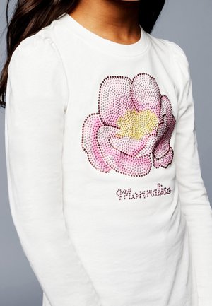 Gros plan d'un enfant portant une chemise blanche à manches longues avec un motif de fleur en strass rose et jaune à l'avant.