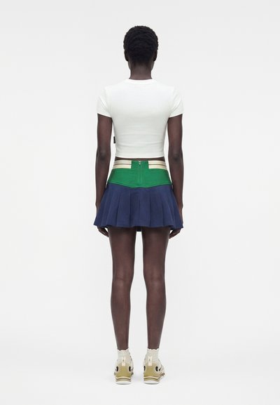 Άτομο που στέκεται με την πλάτη, φορώντας λευκό εφαρμοστό τοπ με κοντά μανίκια, απόχρωσης μπλοκ πράσινη και ναυτική πλισέ φούστα και μπεζ αθλητικά παπούτσια με λευκές κάλτσες.