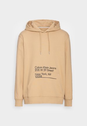 Sweat à capuche beige avec poche avant, capuche à cordon et texte noir au devant indiquant « Calvin Klein Jeans 205 W 39 Street New York, NY 10018 ».