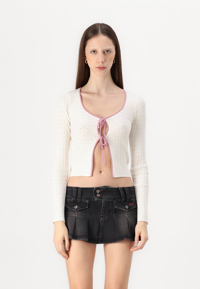 Vit ribbad kort cardigan med rosa detaljer och knytning, i kombination med en svart denim minikjol med knapprader och fickor.