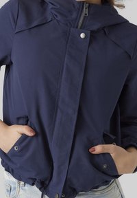 Navyblå lättviktsjacka med huva, framdragkedja, tryckknappsstängning och två sidofickor. Slät textur och elastiska ärmslut.