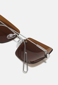 Braun getönte Sonnenbrille mit eckigen Gläsern, metallic-silbernem Rahmen, Nasenpolstern und einzigartigem Doppelschutzdesign. Glatte Oberfläche mit minimalem Zubehör.