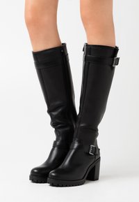 Botas de cuero negras hasta la rodilla con tacones cuadradros, cremalleras laterales y correas con hebillas, llevadas sobre piernas al desnudo contra un fondo blanco sencillo.