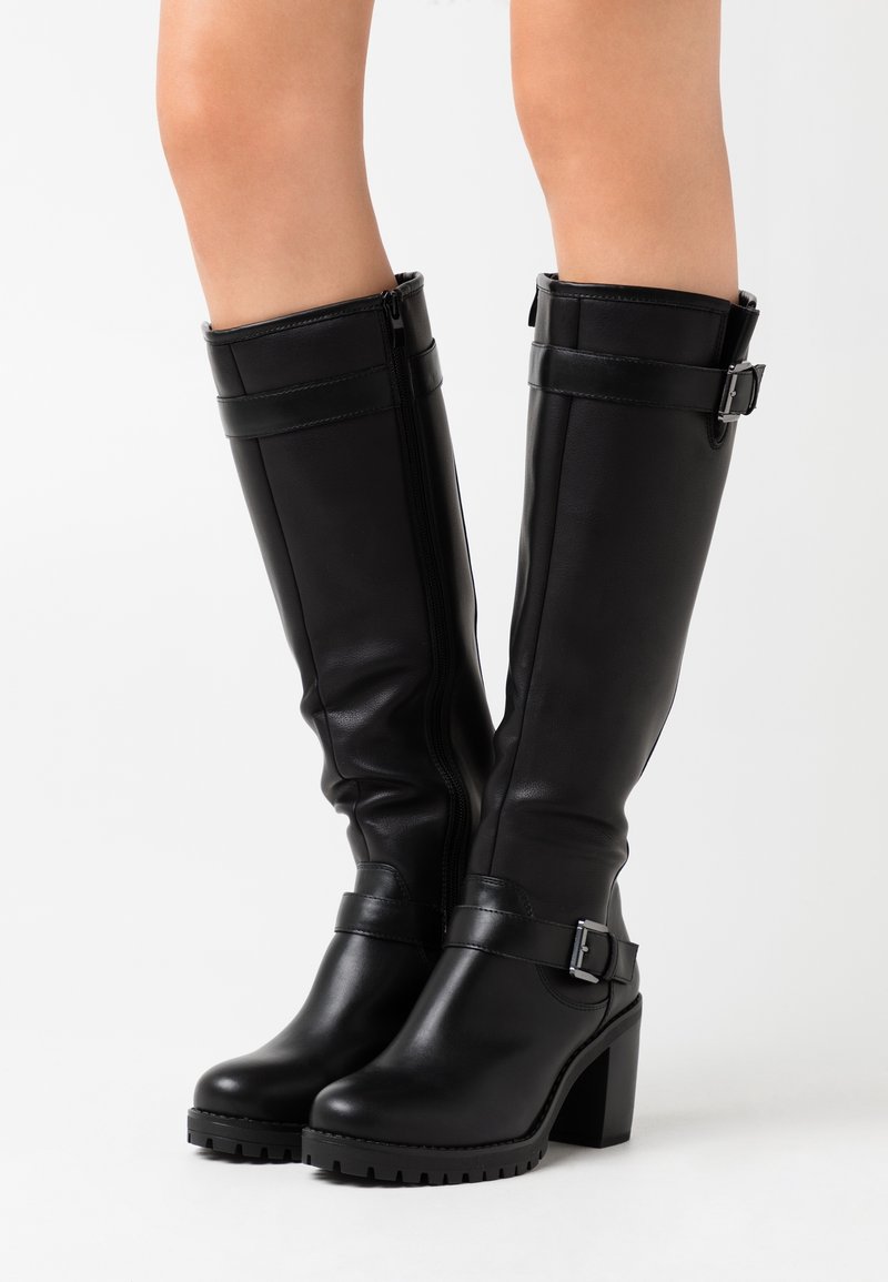 Botas de cuero negras hasta la rodilla con tacones cuadradros, cremalleras laterales y correas con hebillas, llevadas sobre piernas al desnudo contra un fondo blanco sencillo.