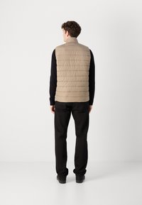 Μπεζ puffer γιλέκο με οριζόντια ραφή, ψηλό γιακά και χωρίς μανίκια. Φοριέται πάνω από μακρυμάνικο μαύρο πουκάμισο και συνδυάζεται με μαύρο παντελόνι.