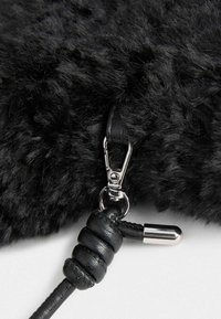 Schwarze Tasche mit strukturiertem Fell, ausgestattet mit einem silbernen Metallreißverschluss, einem schwarzen Ledertragegurt mit einem geknoteten Akzent und einer metallischen Endkappe.