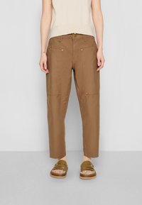 Khaki broek met een relaxte pasvorm, voorzien van zakken aan de voorkant, gouden hardware-accenten en een beschadigde zoom. Gepaard met bruine slide sandalen.