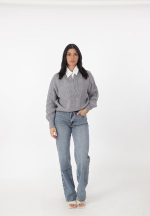 Grijze gebreide trui met een kabelpatroon, gelaagd over een witte overhemddas, gecombineerd met lichtblauwe rechte jeans en sandalen.