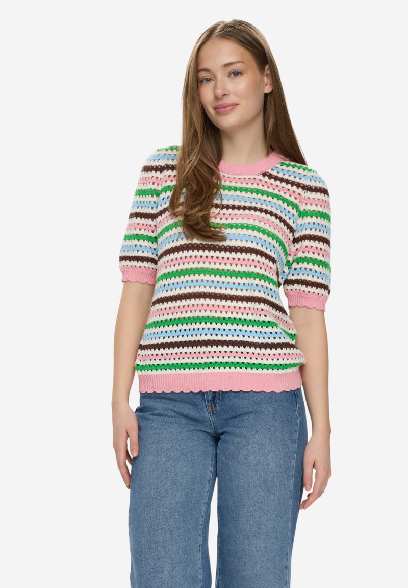 Jeune femme aux longs cheveux portant un pull en maille à manches courtes rayé multicolore et un jean bleu, debout devant un fond uni.