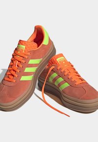 Sneakers featuring a brown suede upper, vibrant orange laces, and lime green accents. Thick sole, three stripes design, and woven tongue detail.