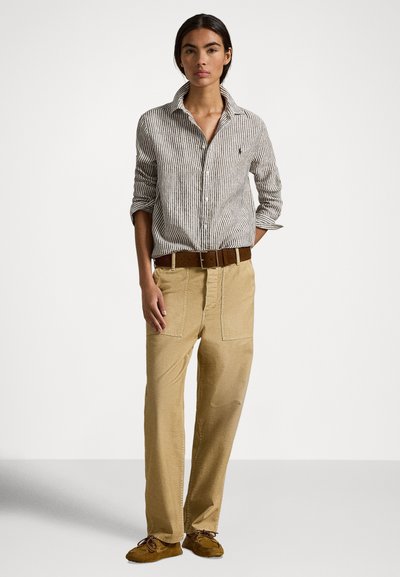 Sieviete valkā svītrainu pogām aizdares kreklu, bēša krāsas plašas bikses ar jostu un brūnas mokasīnu kurpes, stāvot pret vienveidīgu fonu.