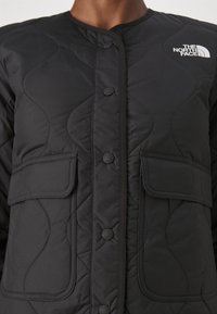 Černá prošívaná bunda s kulatým výstřihem, zapínáním na knoflíky a dvěma kapsami s klopami. Logo je umístěno na horním levém boku.