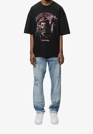 Person iført en sort t-shirt med grafik af en brølende løve, slidte lyseblå jeans og hvide sneakers, stående mod en ensfarvet baggrund.