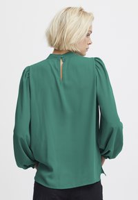 Blusa verde smeraldo con maniche a palloncino, collo rotondo e dettaglio a occhiello sul retro. Realizzata in un tessuto morbido e liscio con un design fluente.