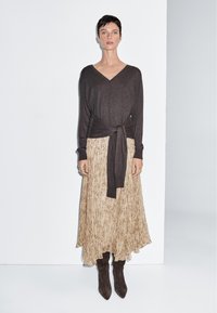 Frau trägt einen dunkelbraunen langärmligen V-Ausschnitt-Pullover mit vorderer Schnürung, beige gemusterten Midirock und dunkelbraune spitze Stiefel, steht vor einer weißen Wand.