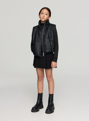 Gilet corto in pelle nera con collo alto e chiusura frontale con zip, abbinato a una maglia aderente nera a maniche lunghe e una gonna nera plisettata. Stivaletti neri alla caviglia.