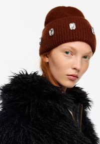 Joven con cabello rojo que lleva un gorro marrón adornado con gemas y un abrigo negro de pelo, mirando hacia la cámara con un fondo blanco.