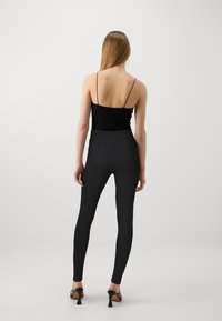Mustavilla pitsillä varustettu bodysuit, jossa on ohuet olkaimet, yhdistettynä korkeavyötäröisiin mustiin leggingseihin. Tasainen rakenne, vartalonmyötäinen siluetti ja nilkkapituus.