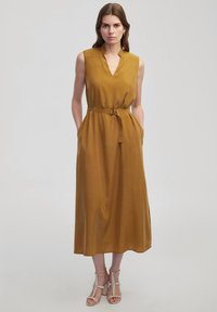 Ärmelloses Senfgelbkleid mit V-Ausschnitt, Gürtel in der Taille, seitlichen Taschen und einer fließenden Silhouette. Glatte Stofftextur.