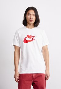 Balta medvilninė marškinėliai su dideliu raudonu ir baltu Nike logotipu ant krūtinės. Dizainas apima trumpas rankoves ir apvalų iškirpimą.