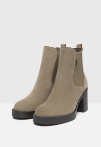 Stivali ankle in suede beige con una finitura liscia, pannelli laterali elastici e un tacco nero spesso. Presentano un anello per una facile calzata.