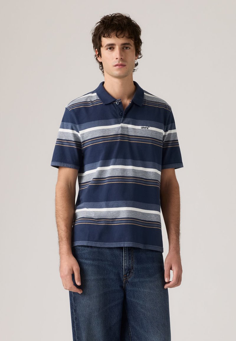 Νέος άνδρας που στέκεται, φοράει μπλε-σκούρο πικέ πόλο Levi's με ρίγες και μπλε τζιν, μπροστά σε απλό ανοιχτό γκρι φόντο.