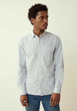 Striped button-up shirt in light blue and white, featuring a chest pocket, long sleeves, and a relaxed fit. Worn with light blue jeans.